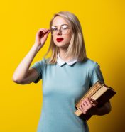 Blonde girl in blue dress and glasses with books on yellow backg Blonde girl in blue dress and glasses with books on yellow background