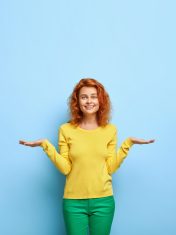 Pleased positive European woman with ginger hairstyle raises two palms as if holding something makes choice between two invisible objects dressed in yellow sweatshirt stands over blue wall blank space Pleased positive European woman with ginger hairstyle raises two palms as if holding something makes choice between two invisible objects dressed in yellow sweatshirt stands over blue wall blank space