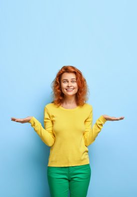 Pleased positive European woman with ginger hairstyle raises two palms as if holding something makes choice between two invisible objects dressed in yellow sweatshirt stands over blue wall blank space Pleased positive European woman with ginger hairstyle raises two palms as if holding something makes choice between two invisible objects dressed in yellow sweatshirt stands over blue wall blank space