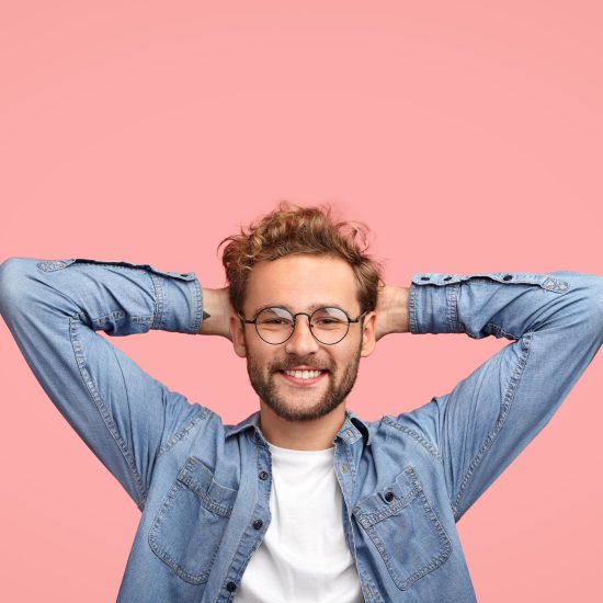 Vertical shot of relaxed carefree man keeps hands behind head, has positive expression, smiles pleasantly, listens something with interest, dressed in fashionable shirt, isolated over pink studio wall Vertical shot of relaxed carefree man keeps hands behind head, has positive expression, smiles pleasantly, listens something with interest, dressed in fashionable shirt, isolated over pink studio wall