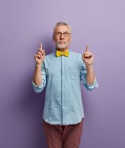 Vertical shot of serious unshaven man of old age, dressed in fashionable clothes, points above, shows blank space for your advertisement, isolated over purple background. Look upside, please Vertical shot of serious unshaven man of old age, dressed in fashionable clothes, points above, shows blank space for your advertisement, isolated over purple background. Look upside, please
